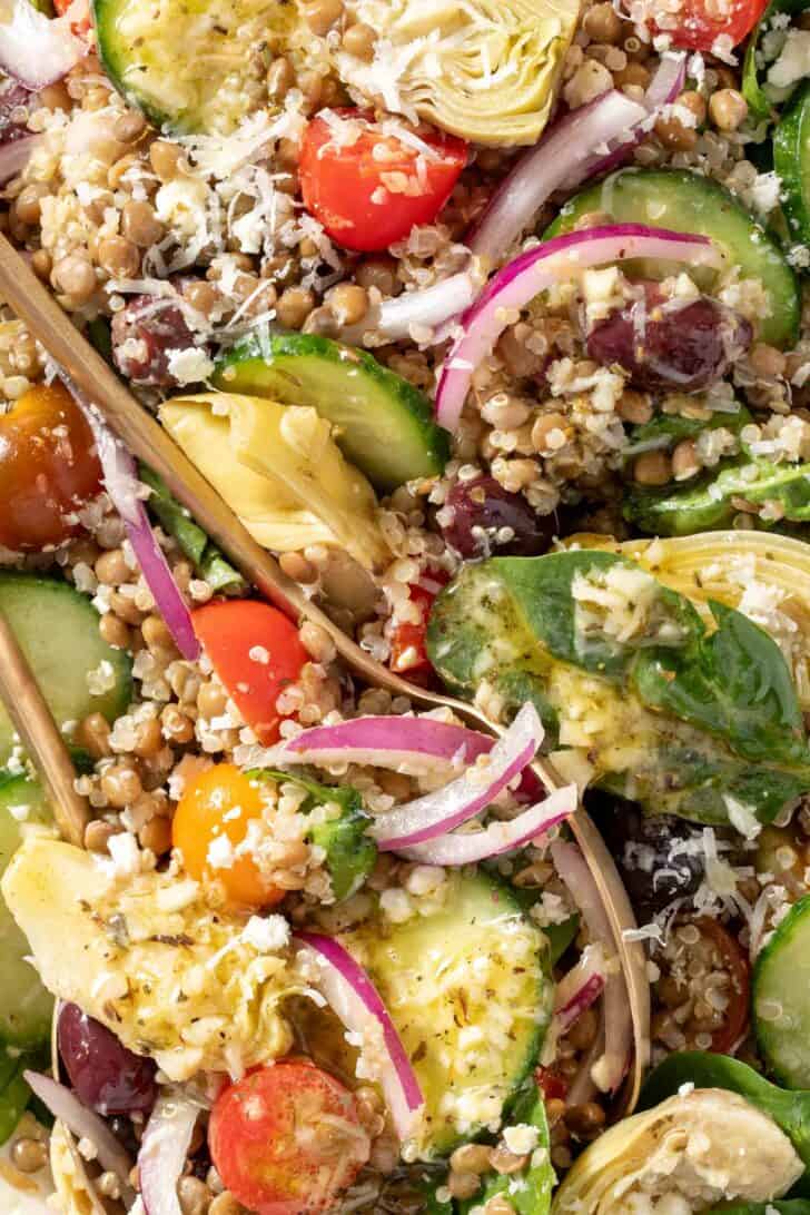 A white platter filled with Lentil Quinoa salad made with lentils, quinoa, spinach, cherry tomatoes, red onion, cucumbers, artichoke hearts and kalamata olives. Gold colored salad servers rest on the platter.