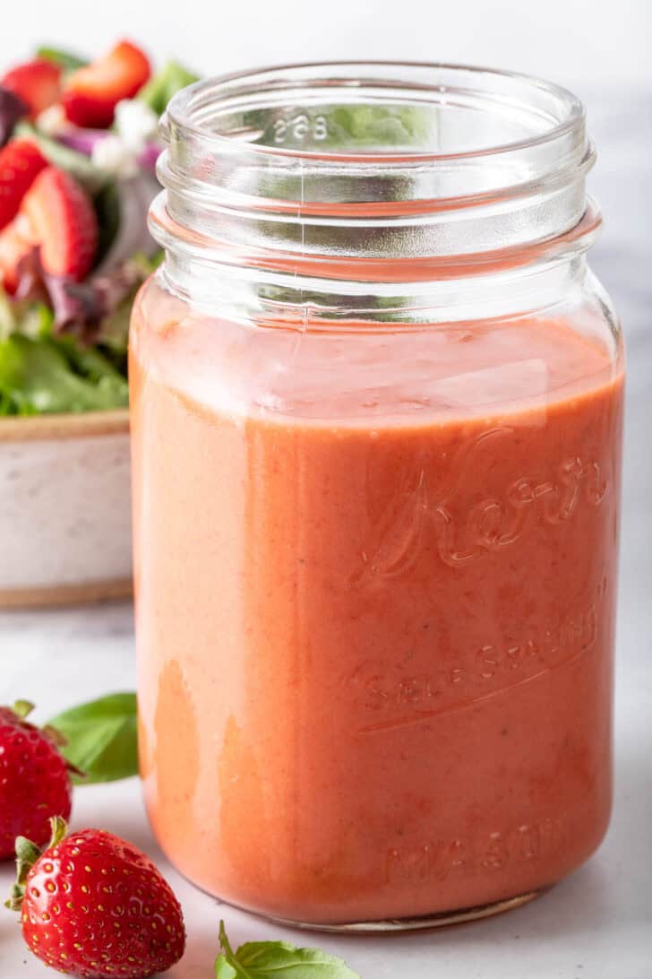 A clear mason jar filled with Summer Strawberry Vinaigrette. A tan colored bowl filled with salad sits next to the jar.