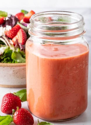 A clear glass mason jar filled with Strawberry Vinaigrette. A tan colored bowl filled with salad sits next to the jar.