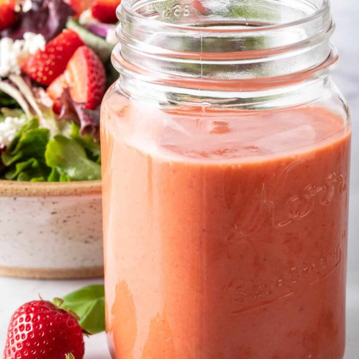 A clear glass mason jar filled with Strawberry Vinaigrette. A tan colored bowl filled with salad sits next to the jar.