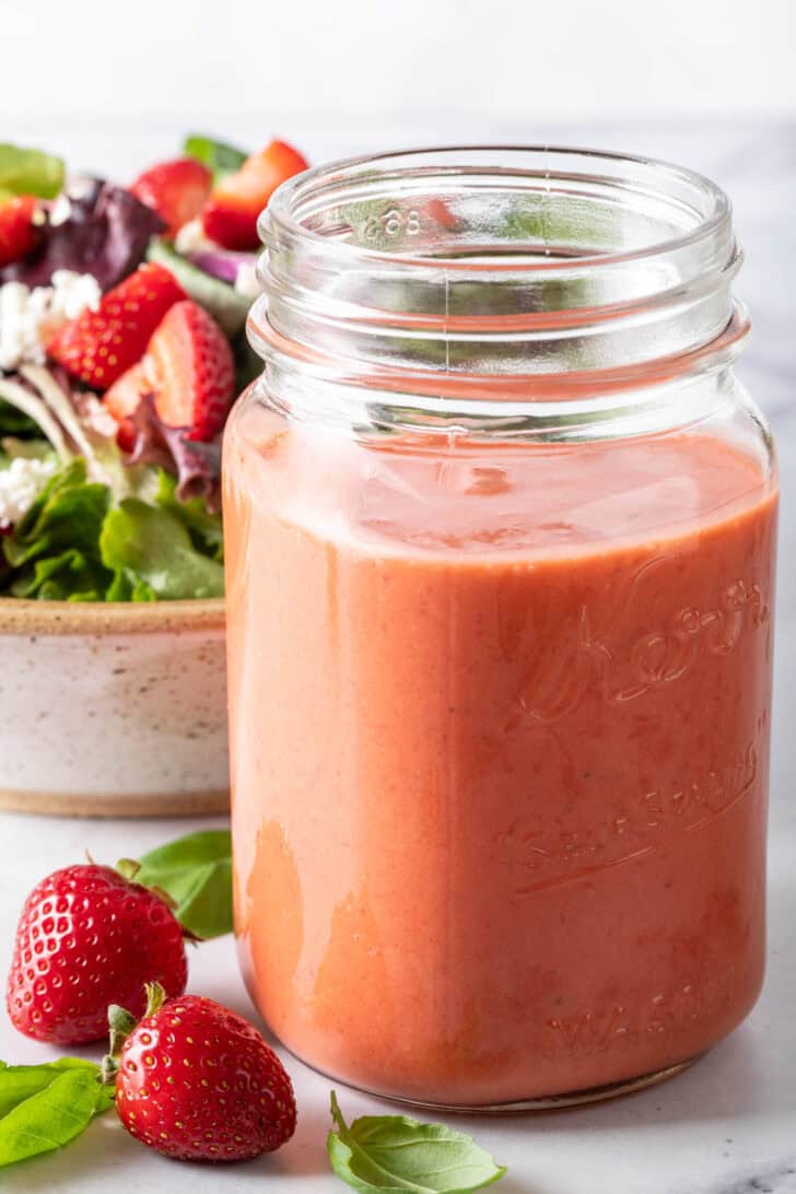 A clear mason jar filled with Summer Strawberry Vinaigrette. A tan colored bowl filled with salad sits next to the jar.