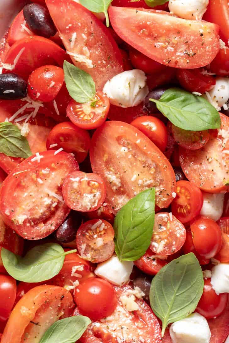 A close up photo of sliced tomatoes with Mozzarella balls kalamata olives and fresh basil leaves drizzled with a vinaigrette.