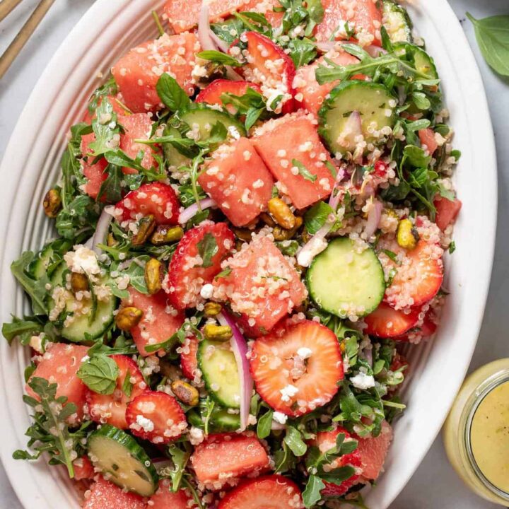 A white oval platter filled with watermelon arugula salad with feta cheese. Gold colored salad tongs sit next to the platter.