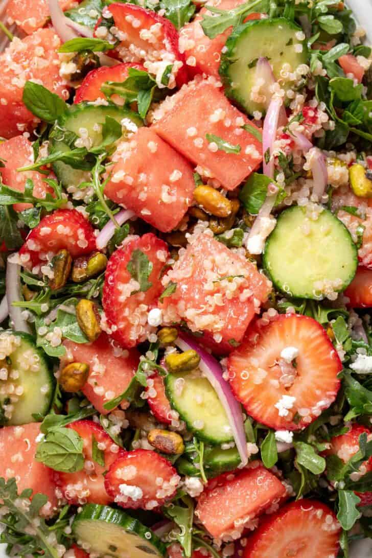 A close up photo of chopped watermelon, slices of strawberry and cucumber tossed with arugula for an Arugula Watermelon Quinoa Salad tossed together.