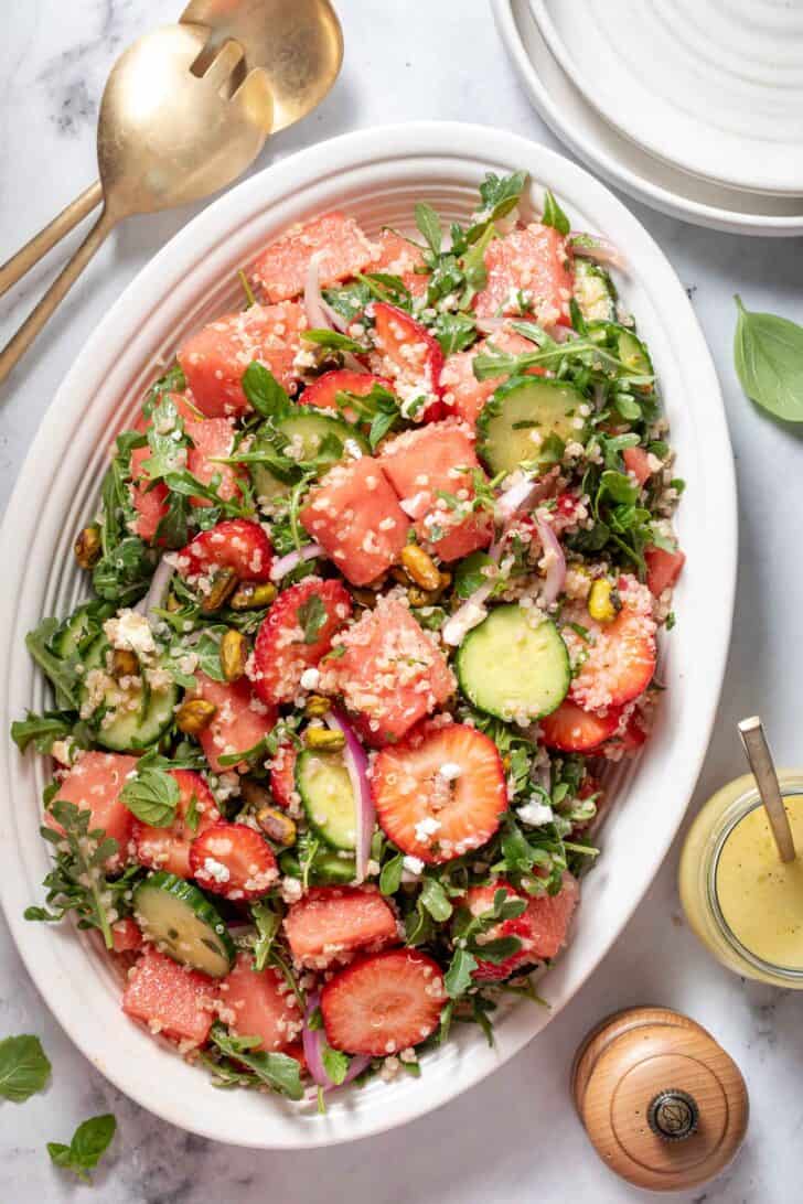 A white oval platter filled with greens and chopped fruit. Gold colored salad tongs sit next to the platter.