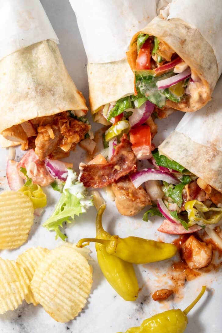 Four BBQ Chicken Wraps laying on a countertop with potato chips scattered next to them.