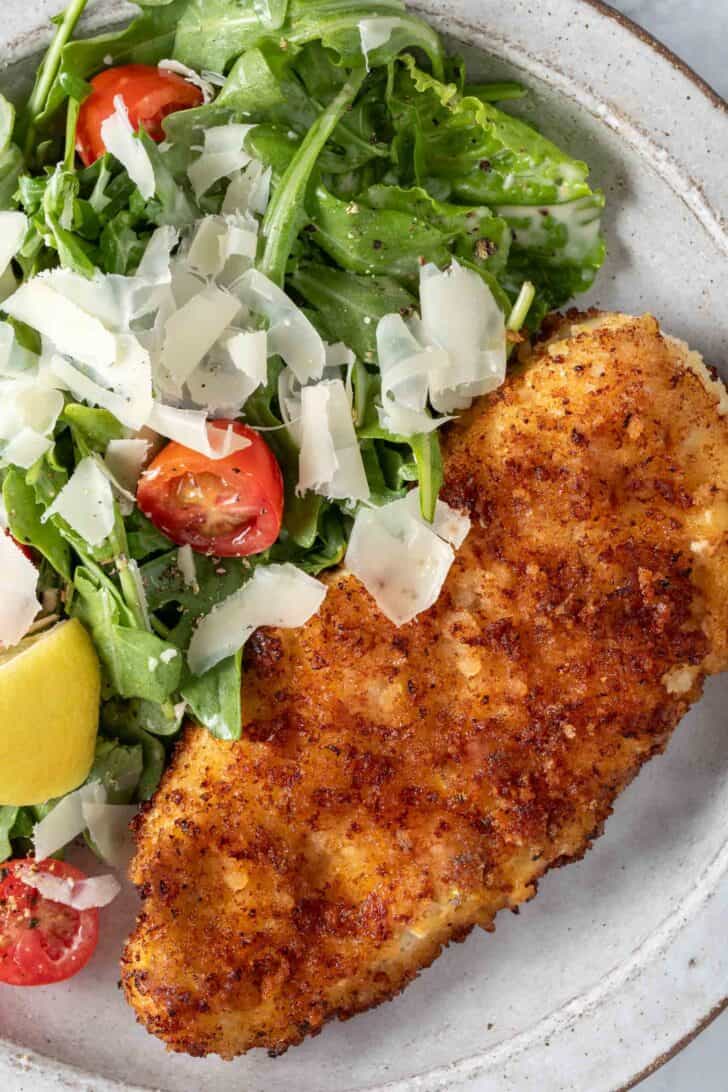 A white plate filled with arugula salad and Parmesan Crusted Chicken cutlet.