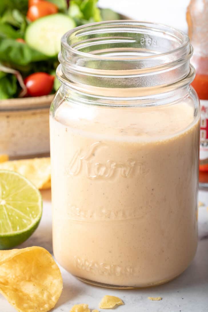 A clear mason jar filled with Buffalo Ranch Dressing. A tan bowl filled with mixed greens sits next to the jar.