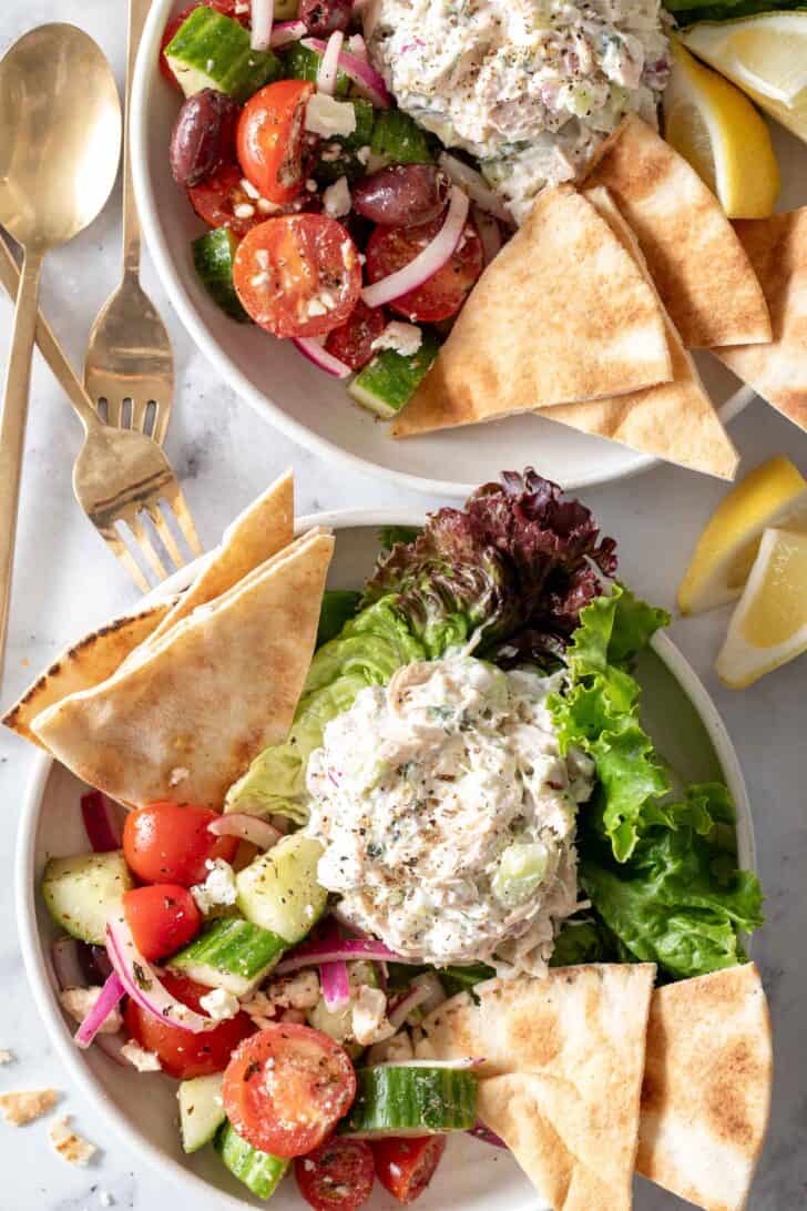 Two white plates filled with scoops of Tzatziki Chicken Salad on lettuce. Pita bread and a cucumber tomato salad sit next to the scoops.