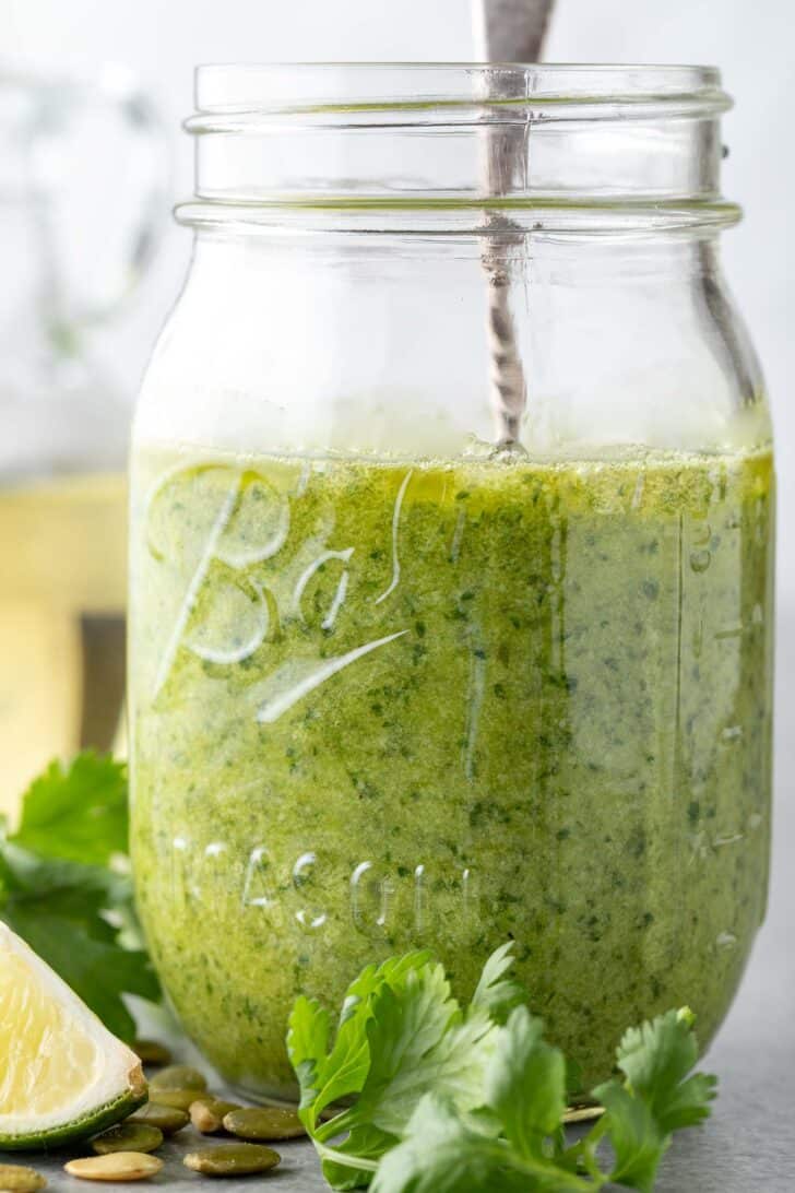 A clear glass mason jar filled with Cilantro Pesto. A spoon rests in the jar.