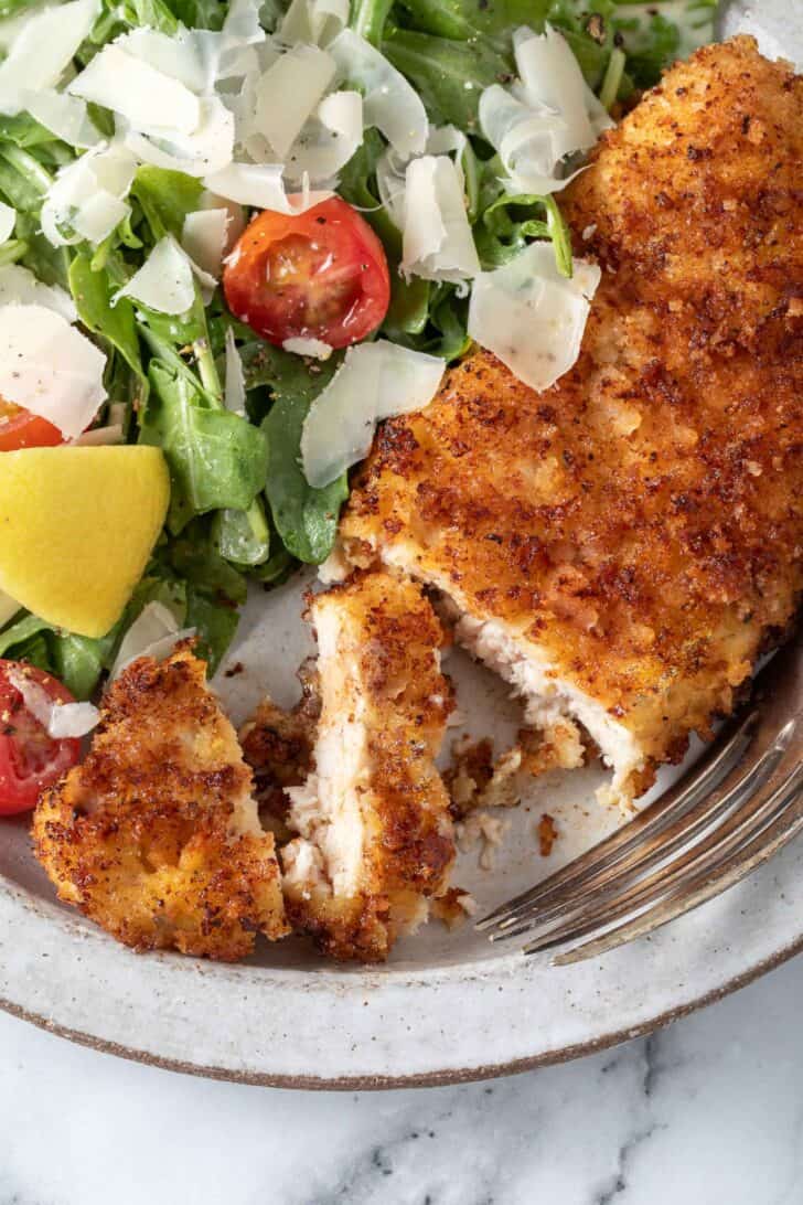 A white plate filled with arugula salad and a Parmesan Crusted Chicken cutlet.