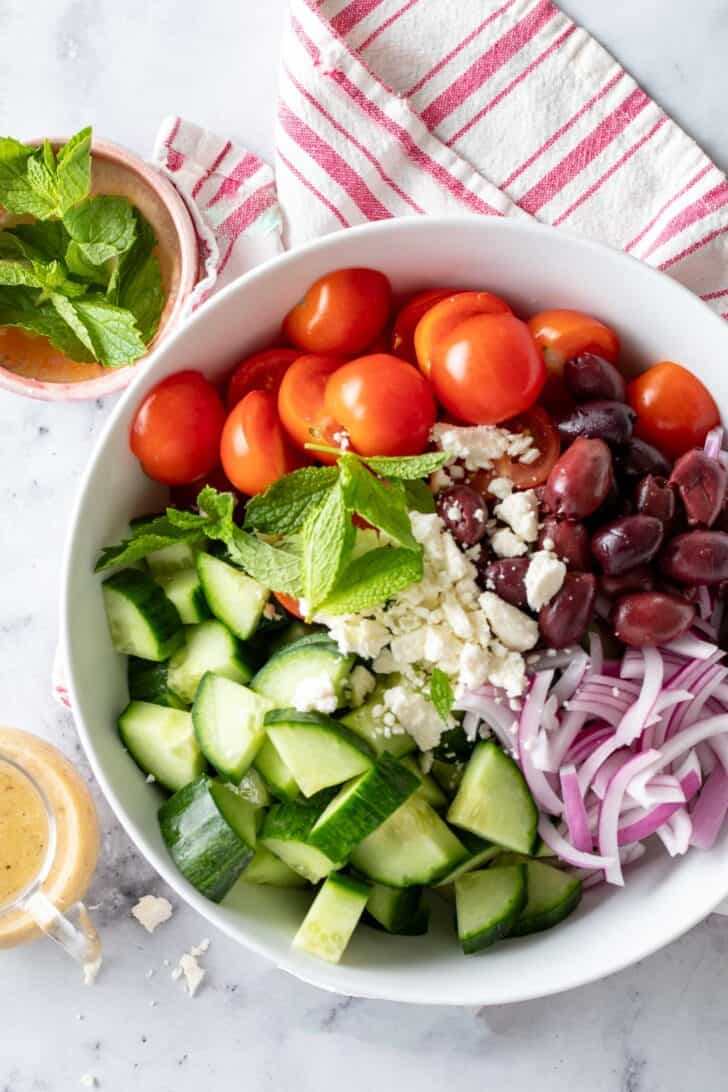 A white bowl filled with chopped tomatoes, olives, vegetables and onions. A small glass is filled with dressing sits next to the bowl.