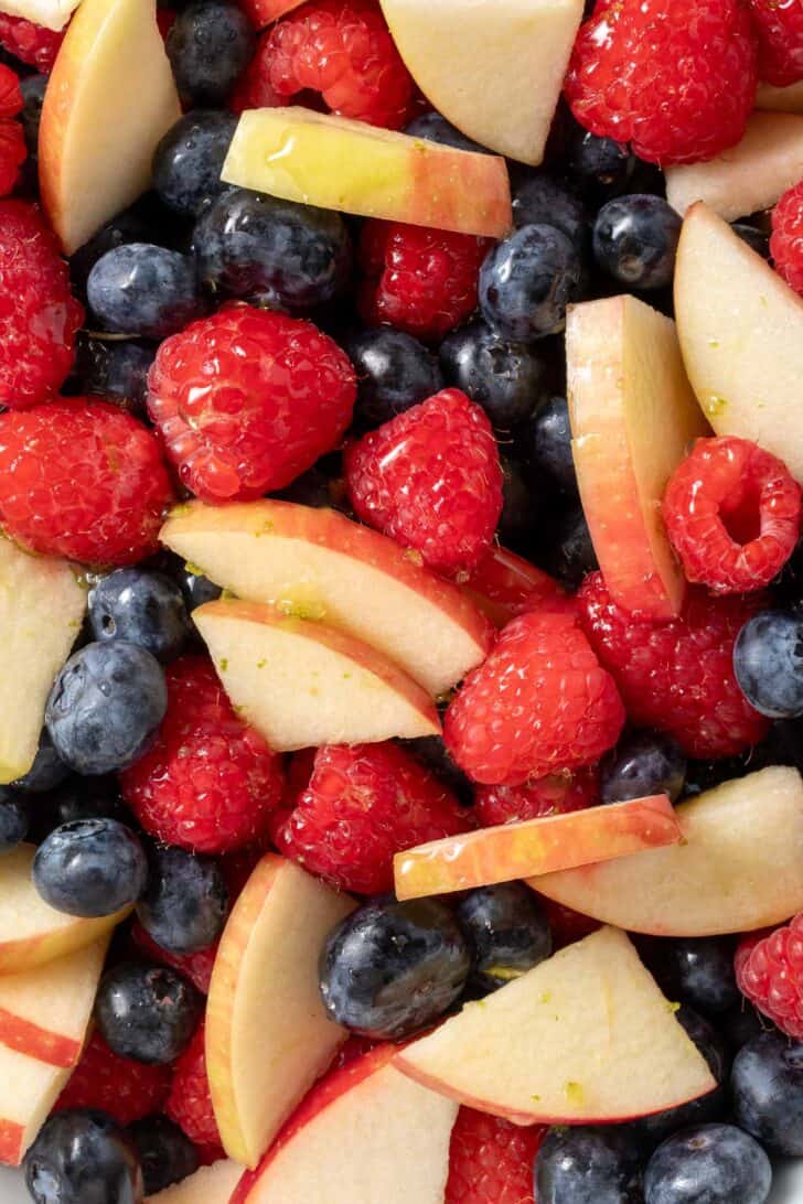 A white bowl filled with raspberries, blueberries and bite size pieces of apple.