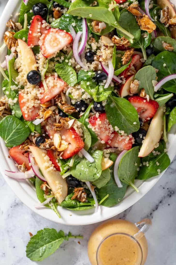 A white platter filled with mixed greens and fruit. A small jar of dressing sits next to the platter.