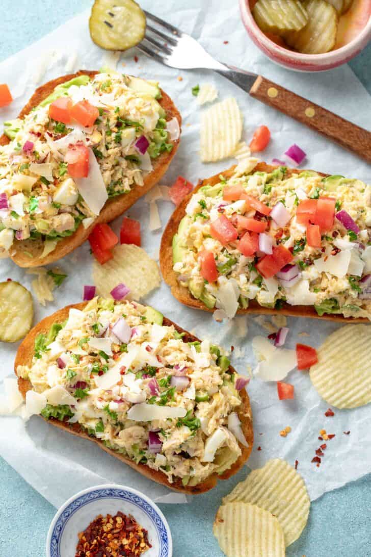 Three open-faced sandwiches on deli paper with potato chips and pickle slices next to them.