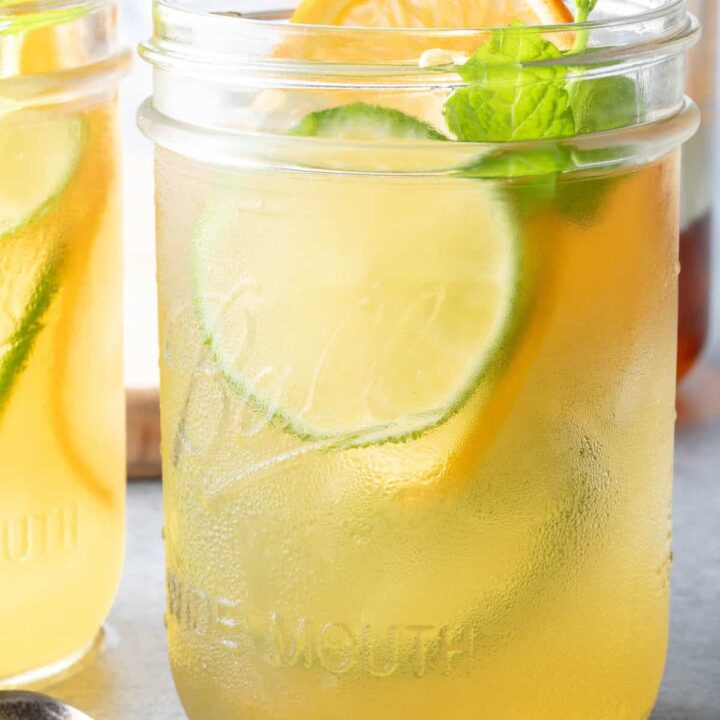 Two clear mason jars filled with Fruit Infused Iced Green Tea. A small spoon filled with honey sits next to the mason jars.