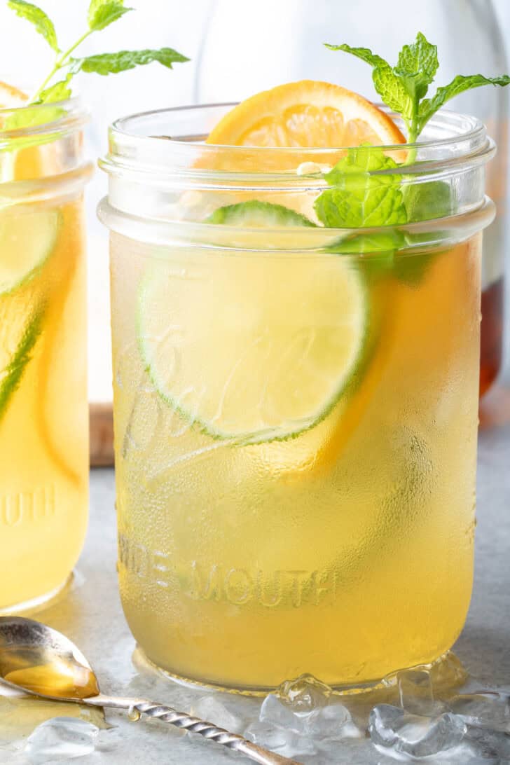 Two clear mason jars filled with Fruit Infused Iced Green Tea. A small spoon filled with honey sits next to the mason jars.