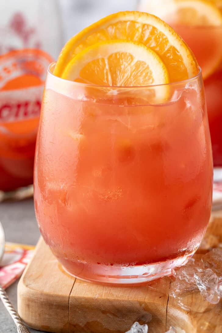 A clear drinking glass filled with Hawaiian Fruit Punch and ice. A lemon and orange slice rest in the drink.