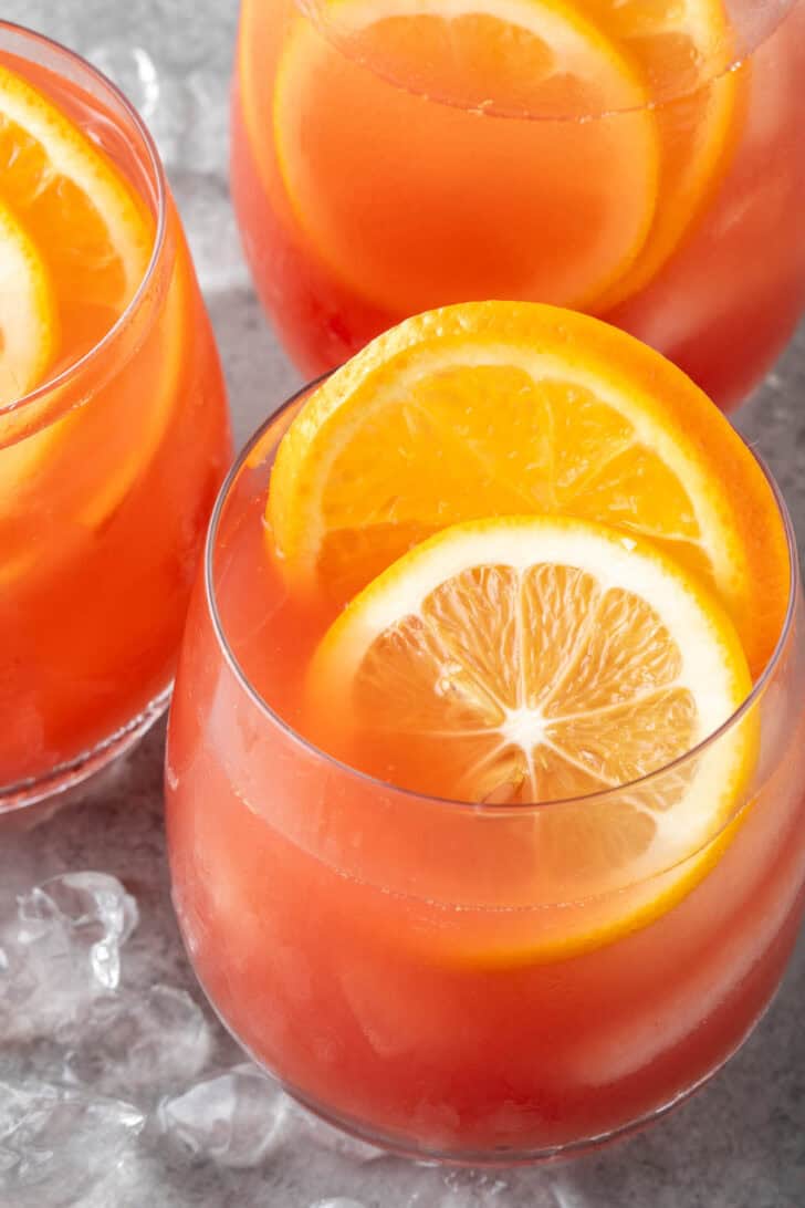 Three clear drinking glasses filled with fruit punch juice and garnished with an orange and lemon slice. Ice is scattered next to the glasses.