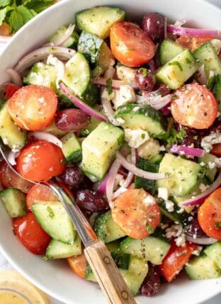A white bowl filled with Greek Cucumber Salad tossed in dressing. A serving spoon with a wooden handle rests in the bowl.