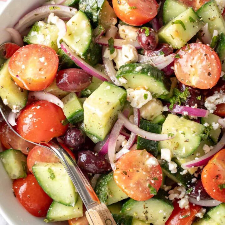 A white bowl filled with Greek Cucumber Salad tossed in dressing. A serving spoon with a wooden handle rests in the bowl.