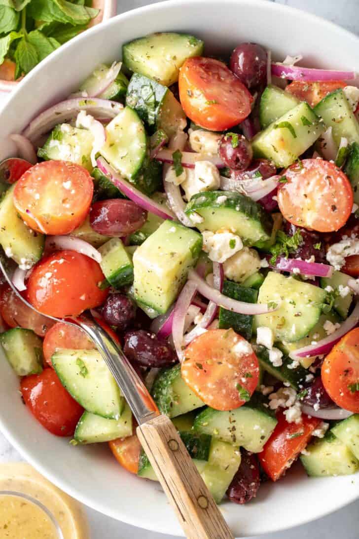 A white bowl filled with Greek Cucumber Salad. A serving spoon with a wooden handle rests in the bowl.