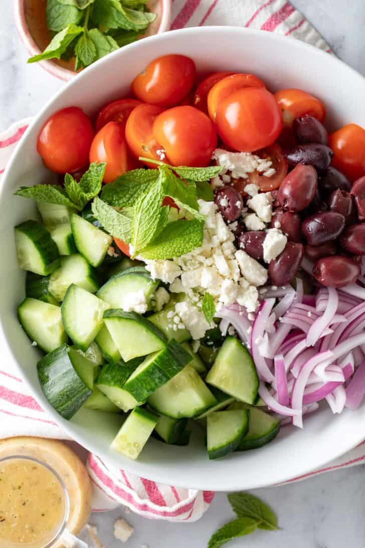 A white bowl filled with tomatoes, olives, sliced onion and vegetables. A small glass filled with dressing sits next to the bowl.