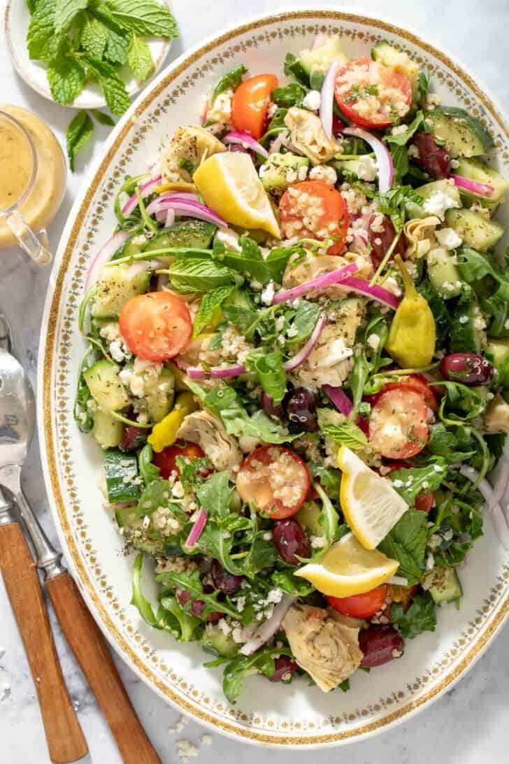 A white oval platter filled with Greek Quinoa Salad. Salad servers with wooden handles rest next to the platter.