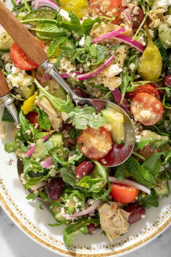 A white oval platter filled with Greek Quinoa Salad. Salad servers with wooden handles rest in the salad.