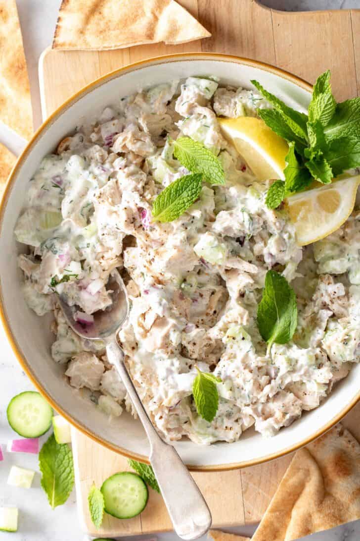 A white bowl filled with chopped poultry tossed in a yogurt sauce. Lemon wedges, and fresh mint leaves are scattered in the bowl.