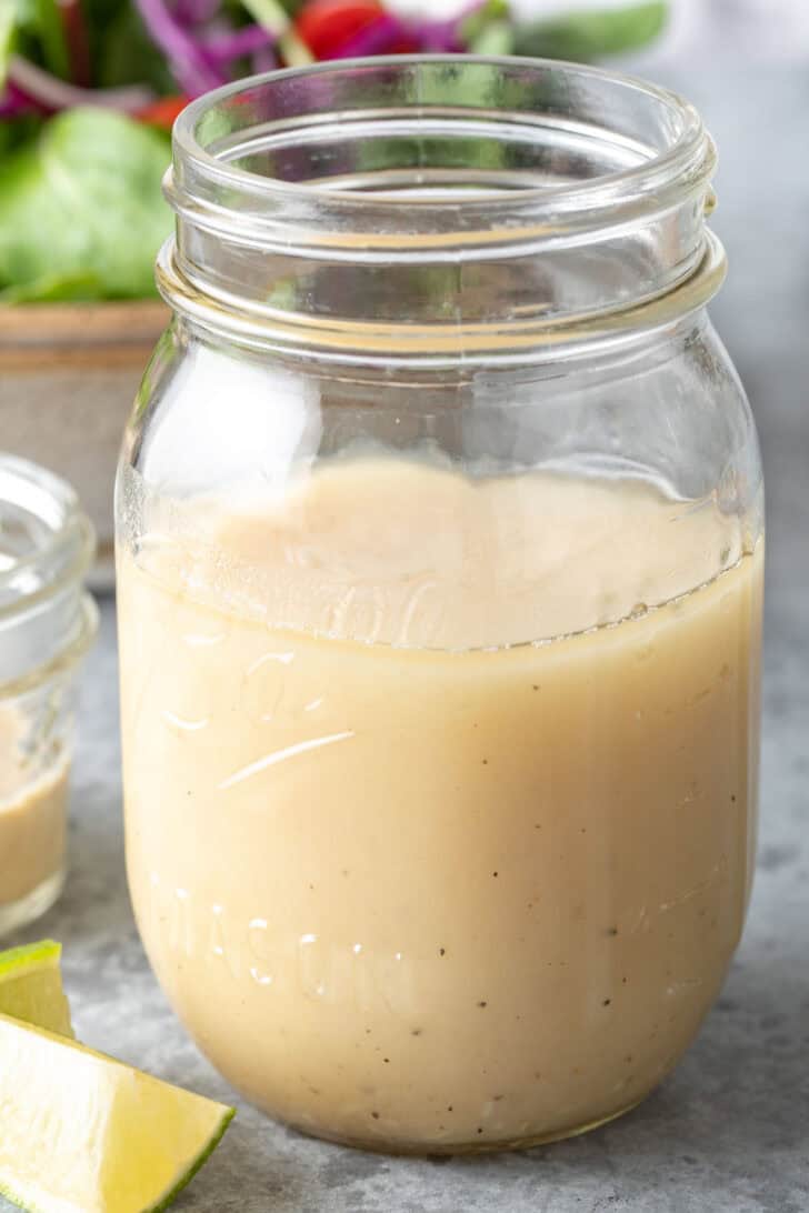 A clear glass mason jar filled with Honey Lime Tahini Dressing. A tan colored bowl filled with salad sits next to the jar.