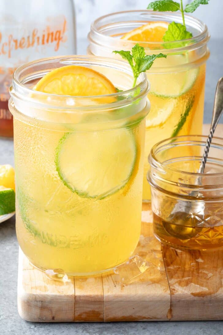 Two clear glass mason jars filled with iced green tea and citrus slices sit on a wood cutting board.