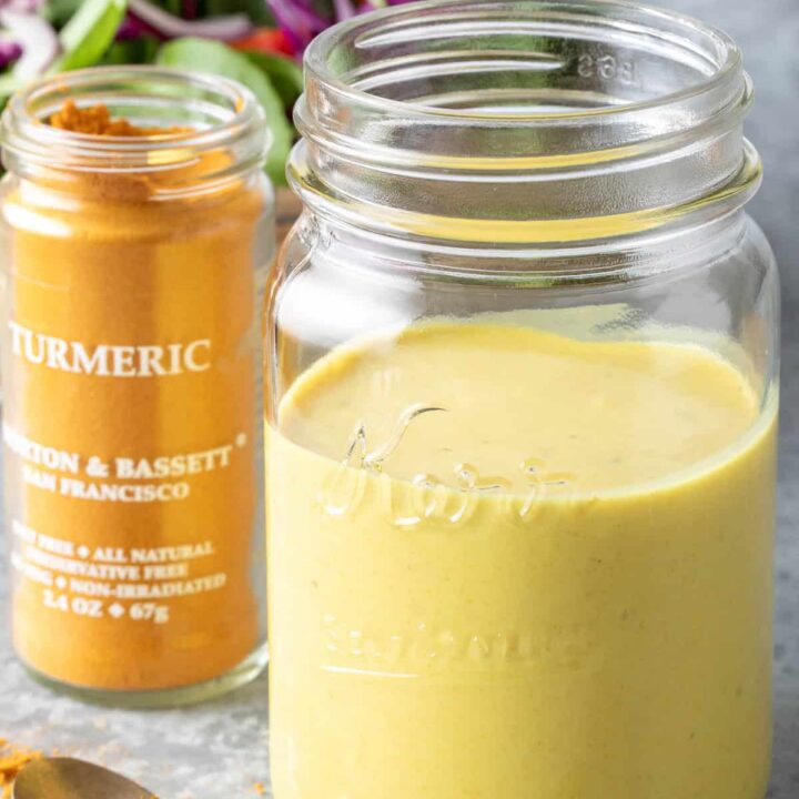 A clear glass mason jar filled with Turmeric Lemon Tahini Dressing. A tan bowl filled with salad sits next to the jar.