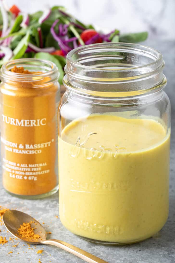 A clear glass mason jar filled with Turmeric Lemon Tahini Dressing. A glass jar with turmeric sits next to the mason jar.