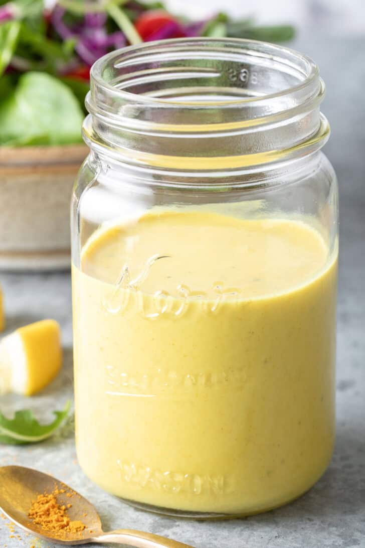 A clear glass mason jar filled with Turmeric Dressing. A tan colored bowl filled with salad sits next to the jar.