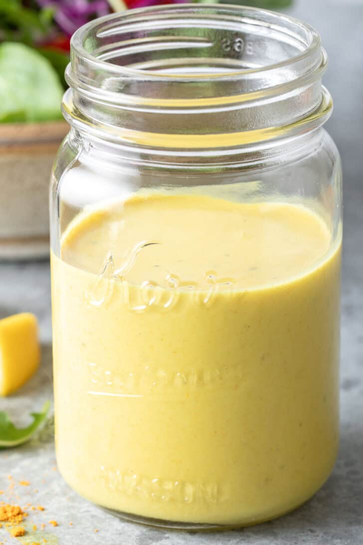 A clear glass mason jar filled with Turmeric Lemon Tahini Dressing. A tan bowl filled with salad sits next to the jar.