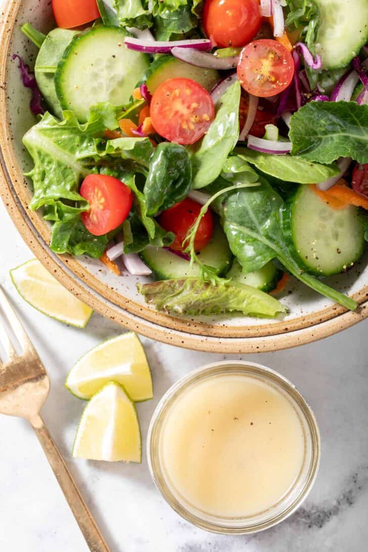 A tan colored bowl filled with green salad. A gold colored fork and a small glass container filled with tahini dressing sits next to the bowl.