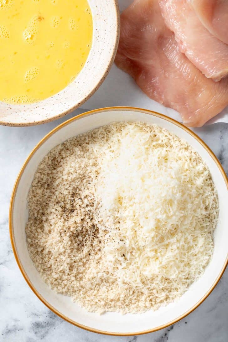 Two white bowls - one filled with bread crumbs and grated Parmesan cheese and the other filled with eggs whisked with milk.