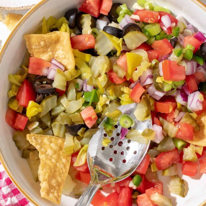 A white bowl filled with Pickle de Gallo made with dill pickles, tomato, onion, garlic. A spoon rests in the bowl.