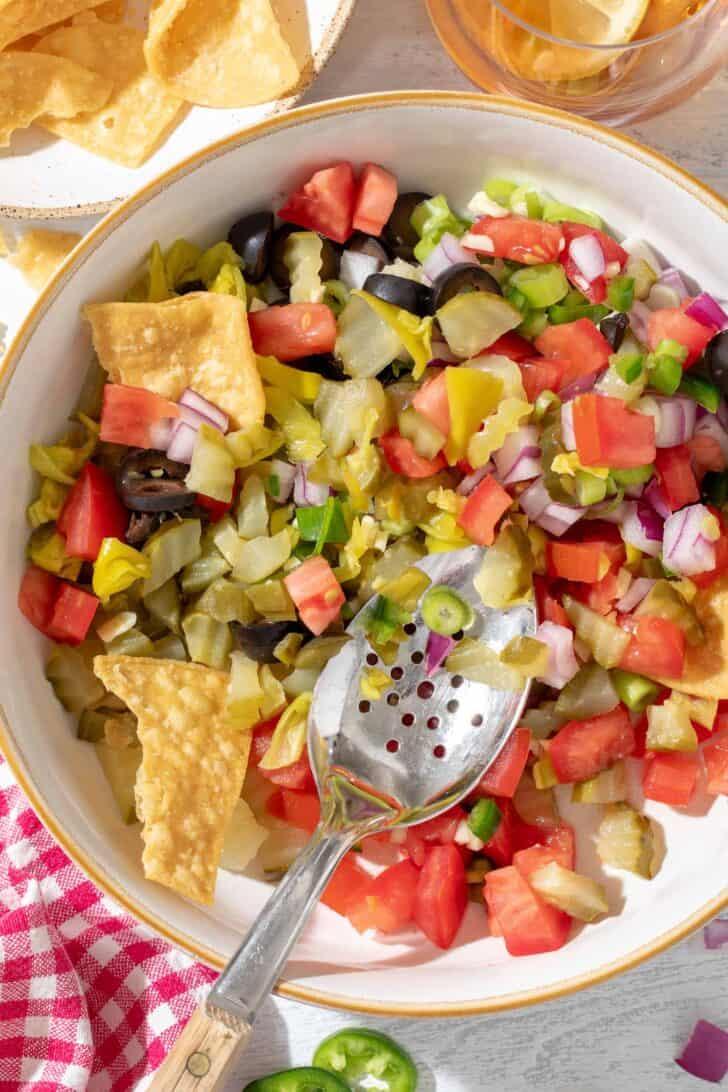 A white bowl filled with Pickle de Gallo made with dill pickles, tomato, onion, garlic. A spoon rests in the bowl.