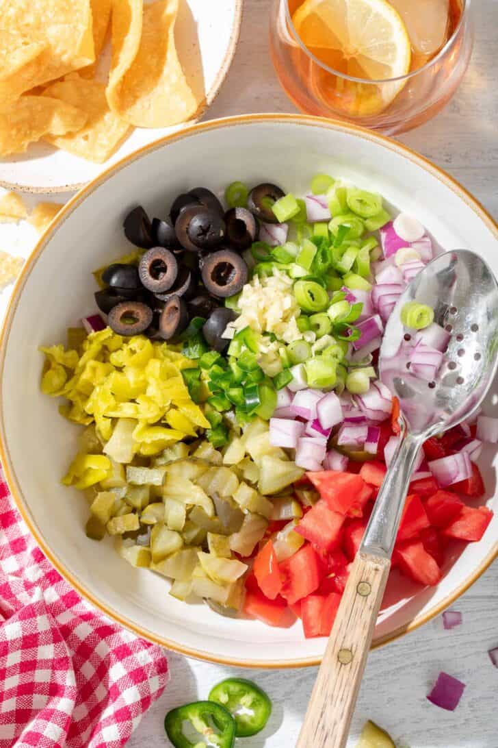 A white bowl filled with chopped black olives, green and red onion, pepperoncini, tomatoes and garlic.