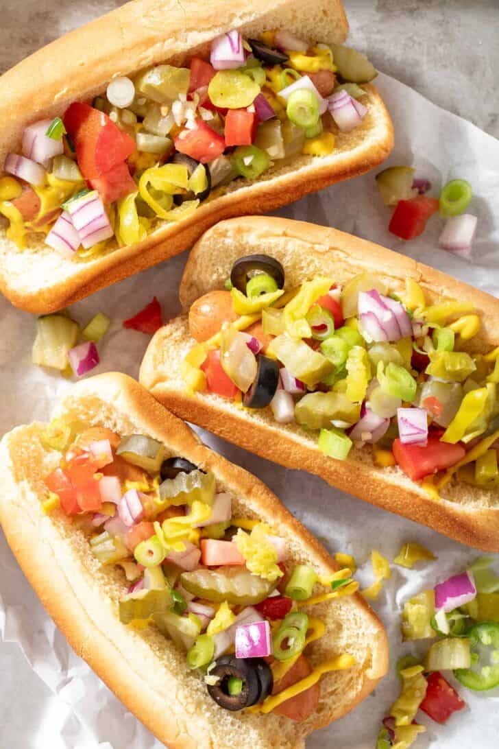 Three hot dogs on buns topped with black olives, pepperoncini, onion, garlic and tomato and mustard.