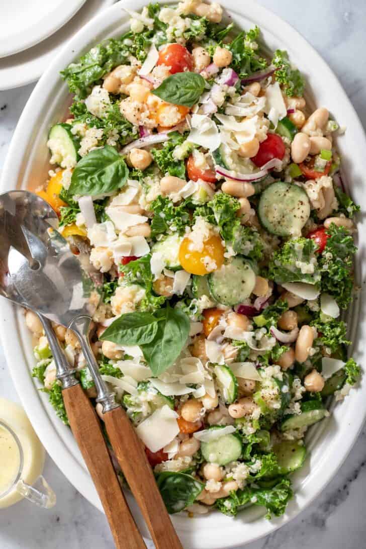 A white platter filled with curly kale, tomatoes, chickpeas and cucumber. Serving spoons with wooden handles rest on the platter.