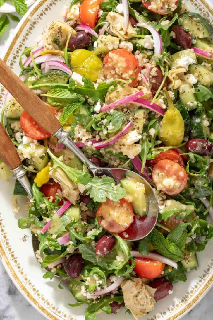 A white oval platter filled with Greek Quinoa Salad. Salad servers with wooden handles rest in the salad.