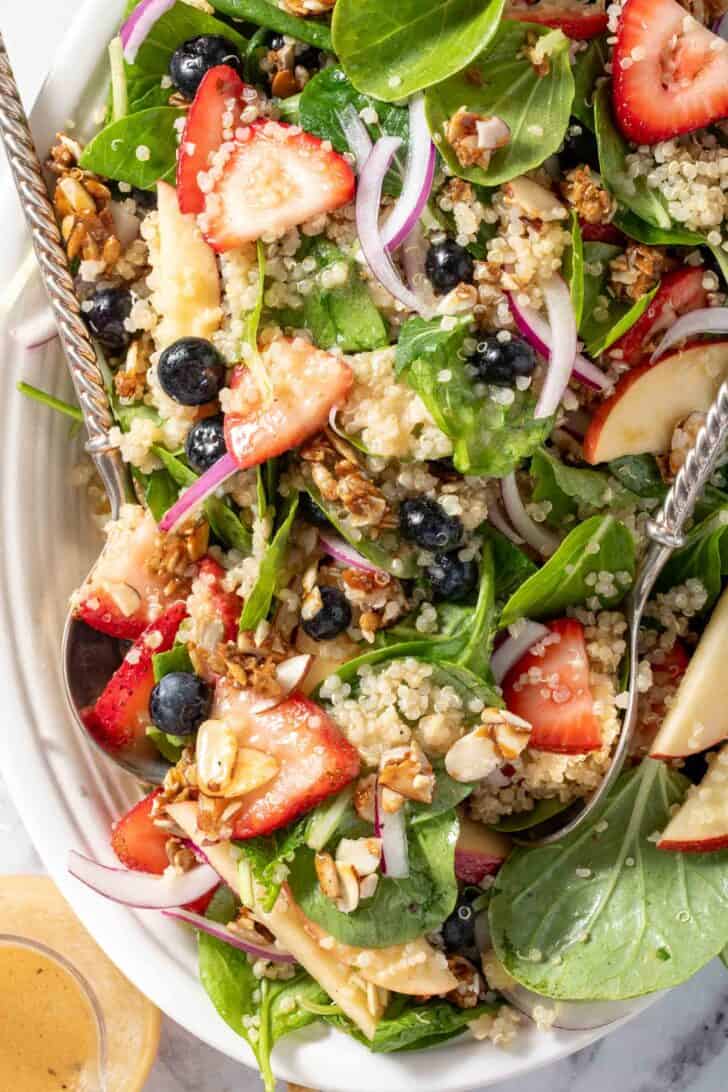 A white platter filled with Strawberry Spinach Quinoa Salad. A pair of silver colored salad servers rest on the platter.