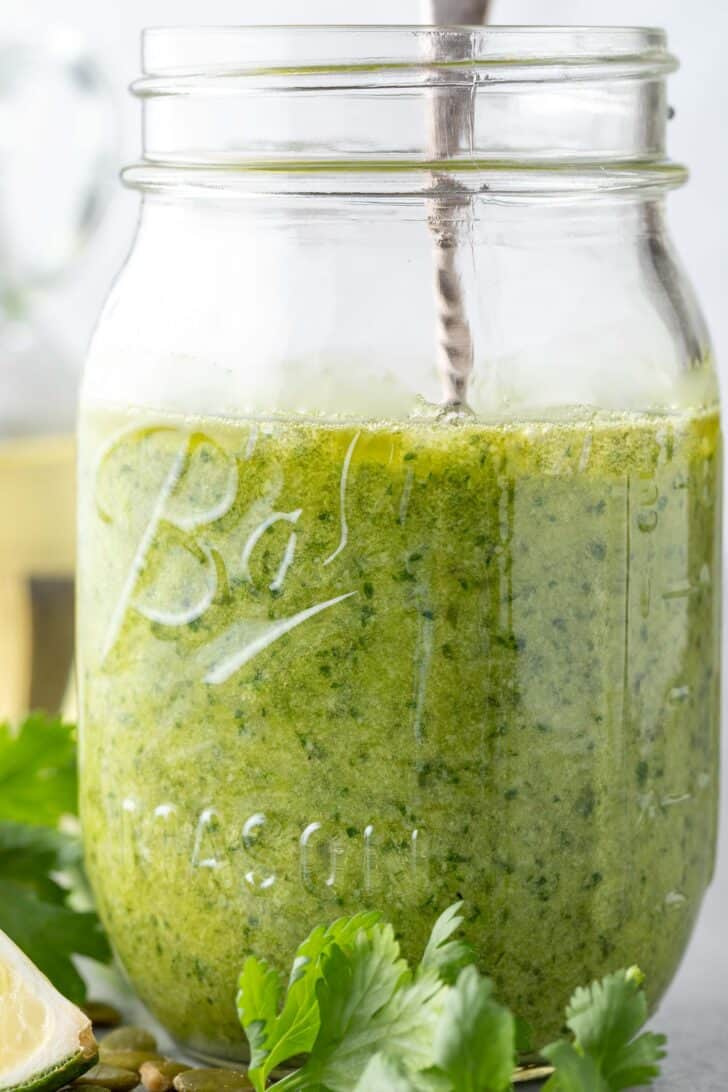 A clear mason jar filled with pumpkin seed cilantro pesto sauce. A silver spoon rests in the jar.