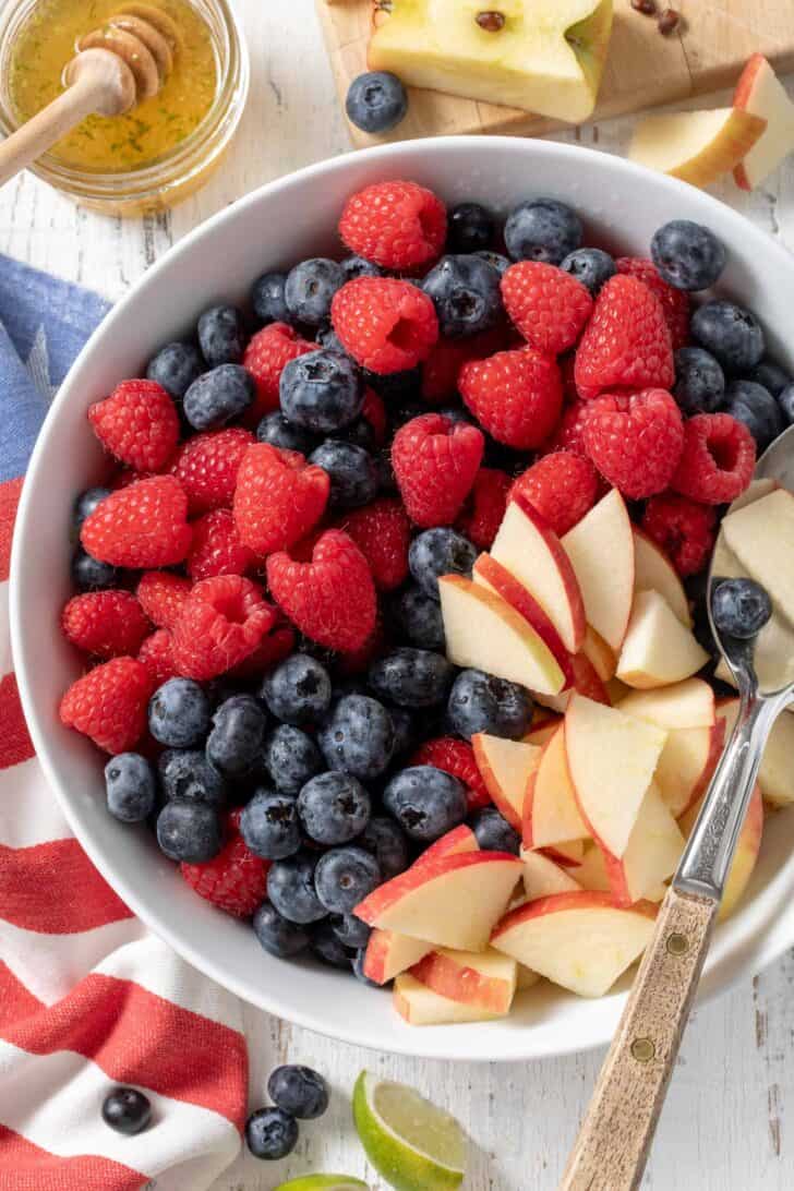 A white bowl filled with raspberries, blueberries and bite size pieces of apple.