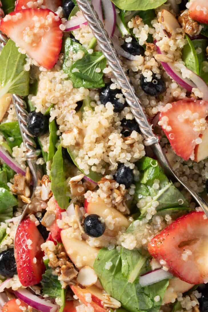 A white platter filled with Strawberry Spinach Quinoa Salad. A pair of silver colored salad servers rest on the platter.