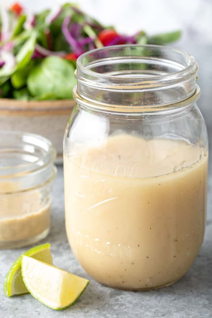 A clear glass mason jar filled with a creamy vinaigrette. A tan colored bowl filled with mixed greens sits next to the jar.