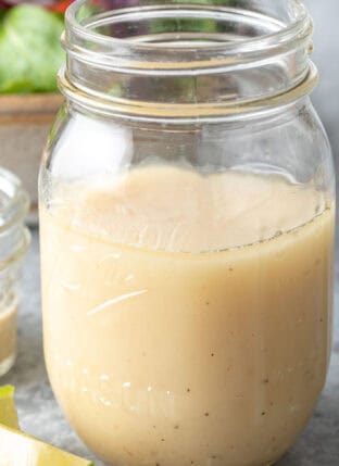 A clear glass mason jar filled with Honey Lime Tahini Dressing. A tan colored bowl filled with salad sits next to the jar.